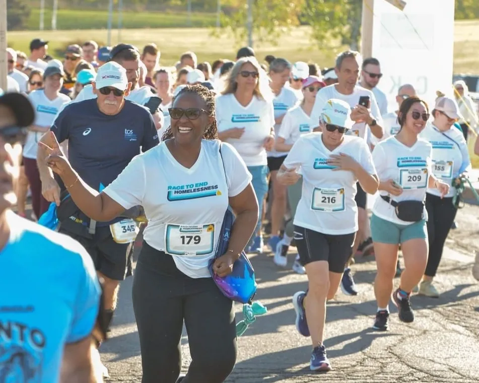 A group of runners in Race to End Homelessness t-shirts participating in a race, with one woman in the foreground smiling and waving at the camera.