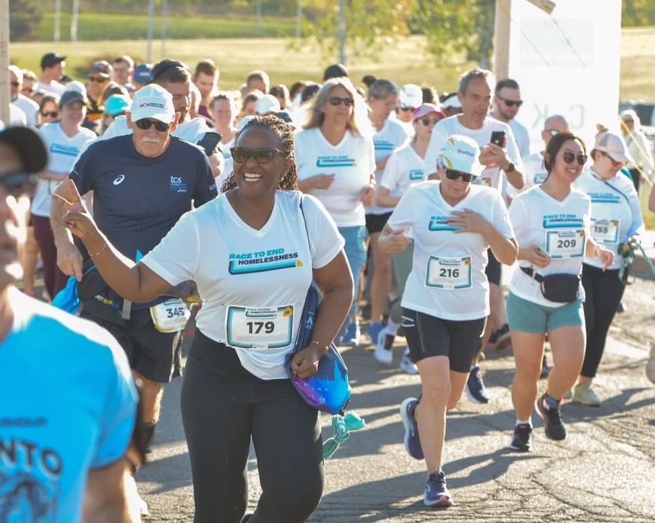 Une femme souriante, portant un dossard, court au milieu d'une foule de personnes vêtues de t-shirts blancs portant l'inscription Race to End Homelessness.