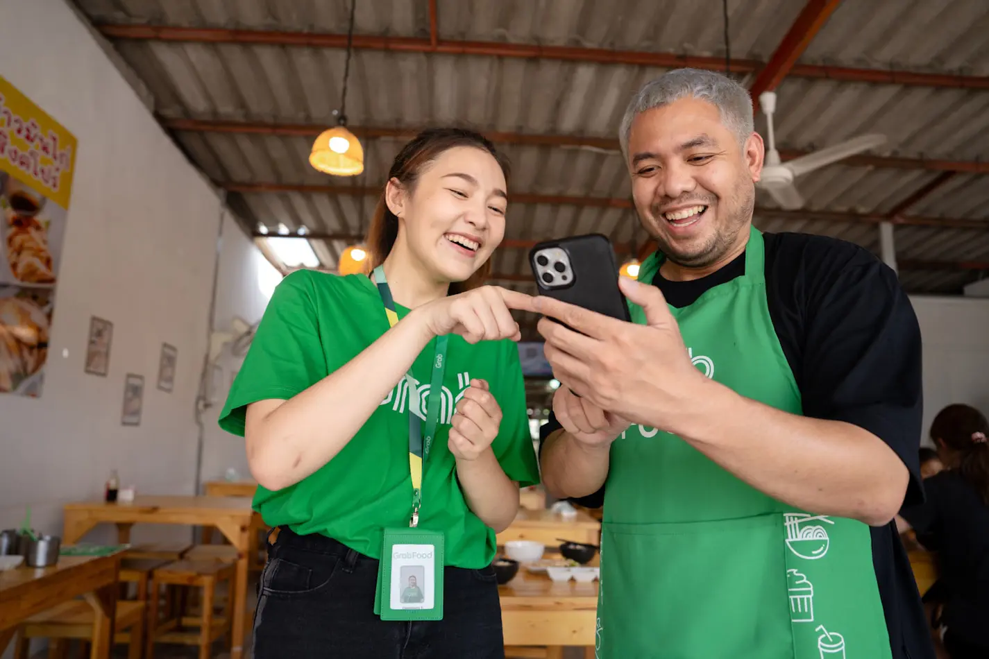 A man and a woman wearing green GrabFood uniforms smile while looking at a smartphone together.