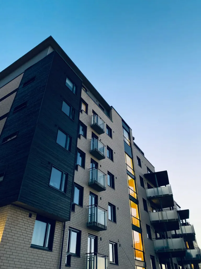 A low-angle view of a modern multi-story apartment building with dark and light-colored facades, glass balconies, and glowing yellow windows against a clear blue sky.