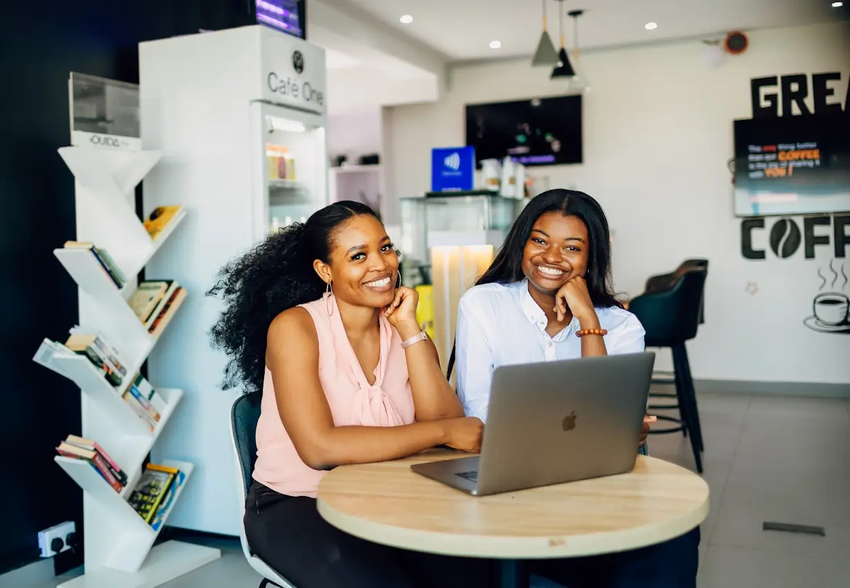 Two smiling women sit at a round table with a laptop in a cafe.