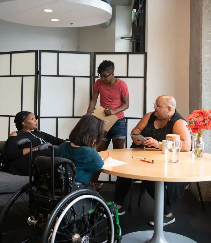 Four people gather around a round table in an office setting, with one person in a wheelchair and another standing while holding a clipboard.