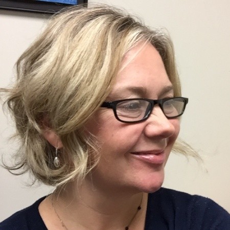 Une femme souriante aux cheveux blonds mi-longs, portant des lunettes à monture noire.