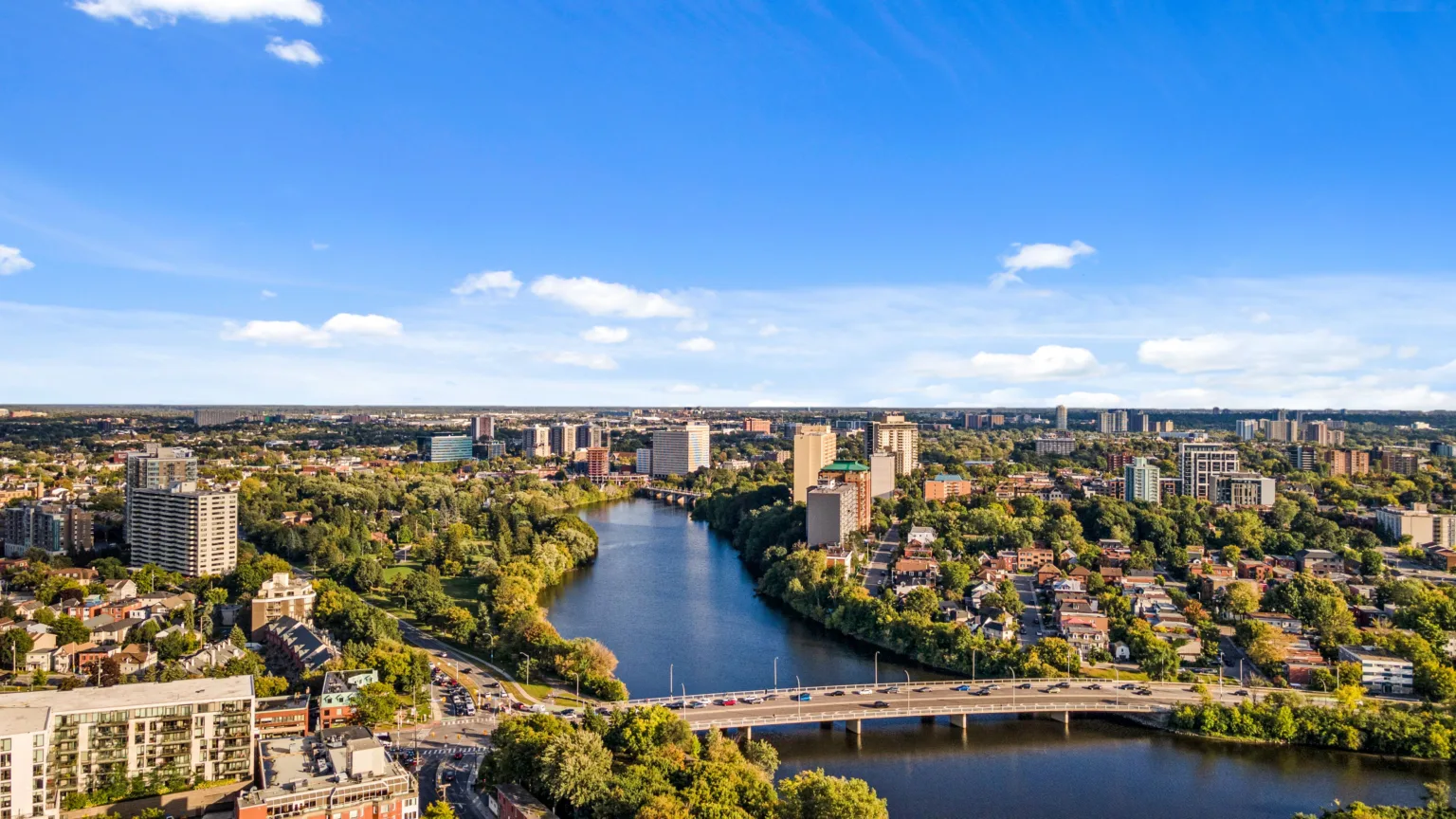 Aerial view of a river flowing through a city with bridges, high-rise buildings, and dense tree cover under a clear blue sky.