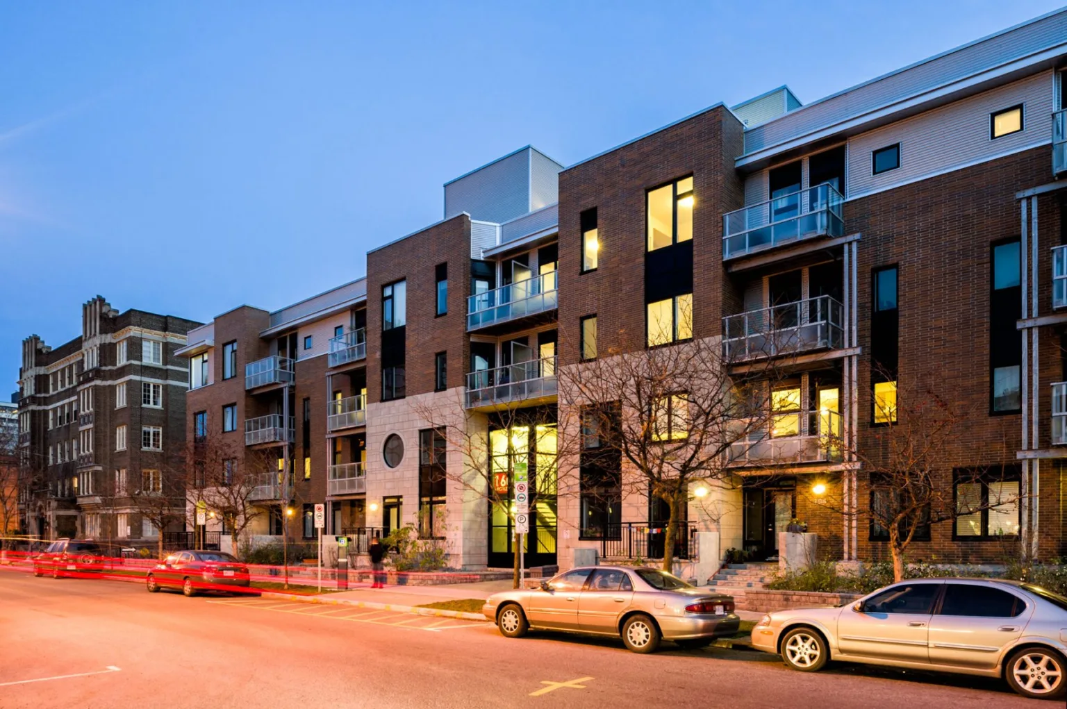 A multi-story brick apartment building at dusk with parked cars on the street and light trails from a moving vehicle.
